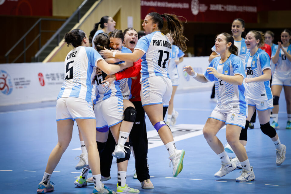 A group of athletes in light blue and white uniforms embrace in celebration on an indoor court, with teammates cheering nearby.